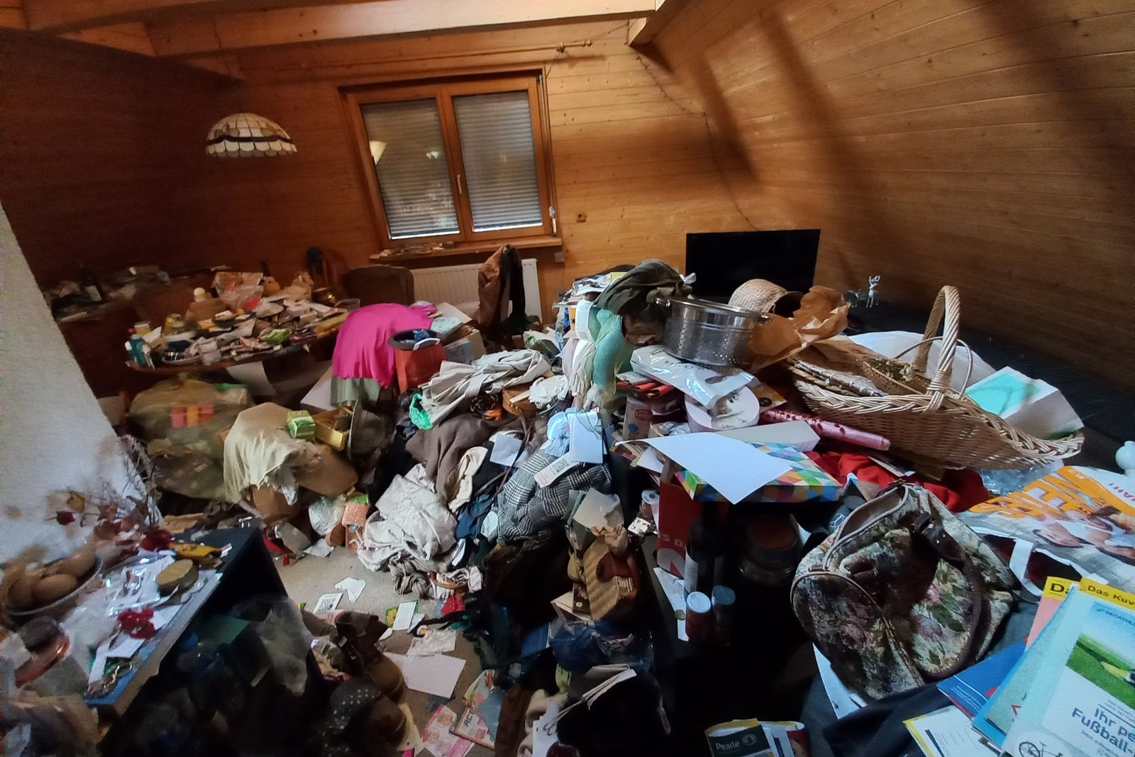 Pièce mansardée lambrissée d'un appartement envahie par un amas de vêtements, sacs, papiers et objets entassés au sol et sur les meubles