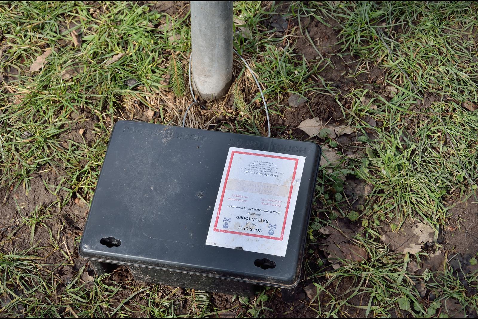 Poste d'appâtage noir contre les rats, fixé par un câble à un poteau et portant une étiquette d'avertissement, posé dans l'herbe d'un parc