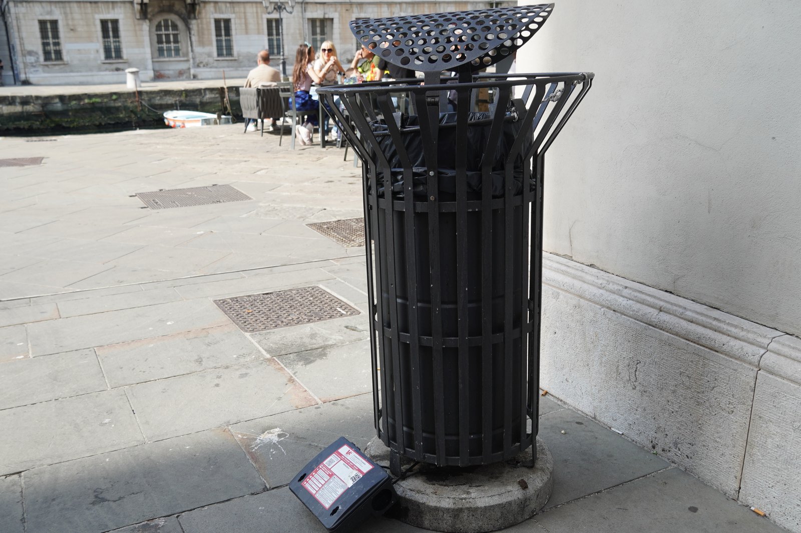 Poste d'appâtage noir contre les rongeurs posé au pied d'une corbeille à déchets métallique, sur une place pavée
