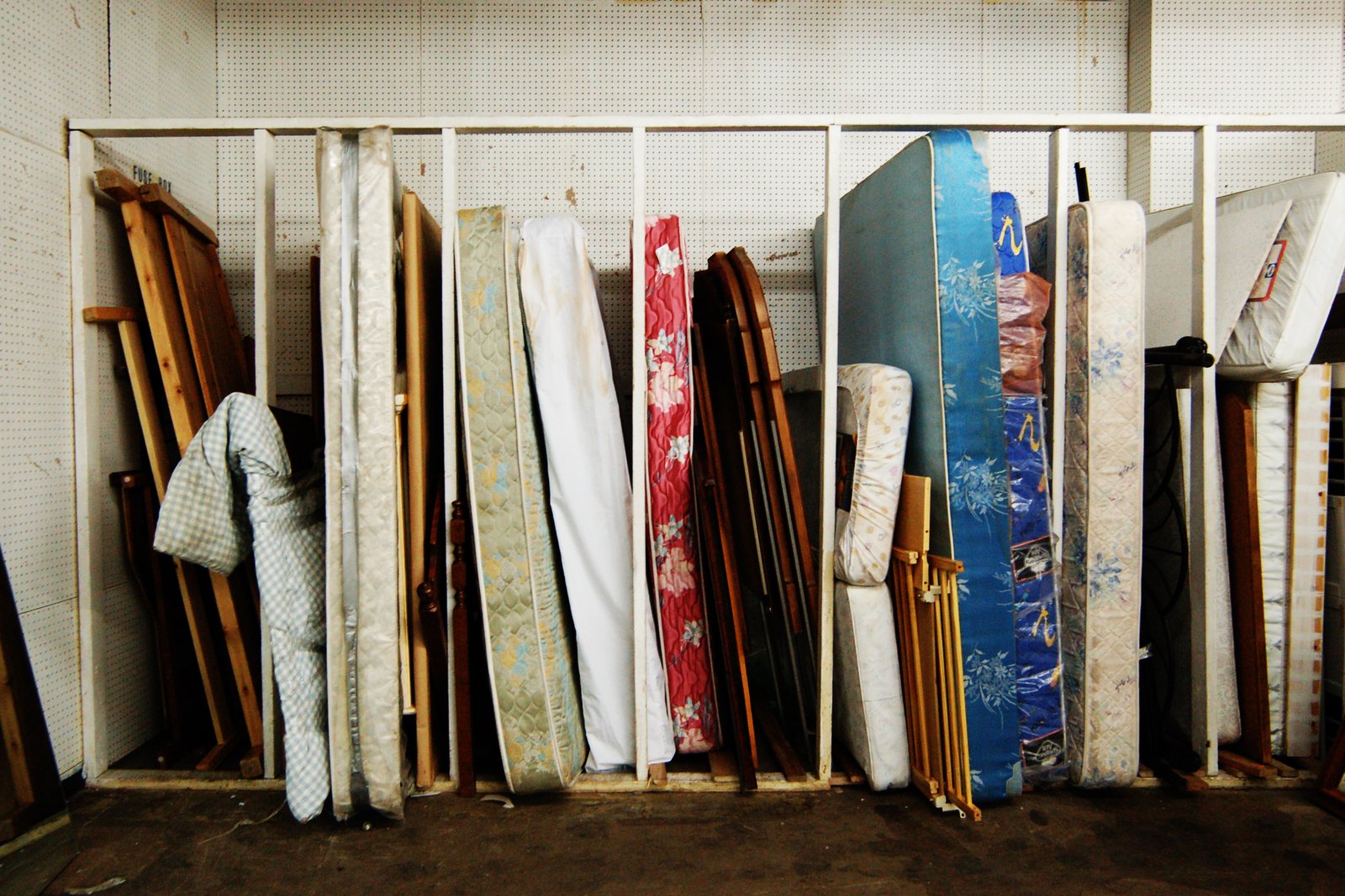 Une rangée de matelas usagés de couleurs différentes et des montants de lit en bois, debout côte à côte contre un mur perforé